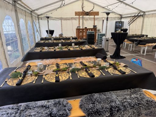 „Stimmungsvolles Festzelt mit dunklen Tafeln, goldenem Glanz, warmer Grüngirlande und einer rustikalen Bar, die alles in ein gemütlich-elega